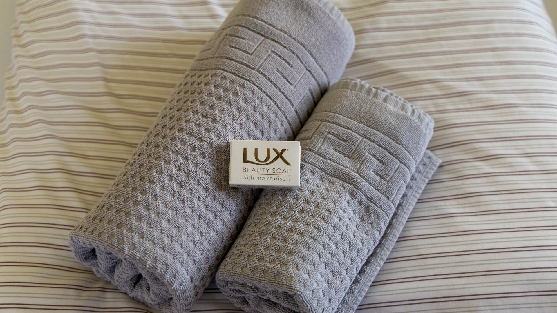 Luxuriöse Handtücher und Seife Zwei graue, gerollte Handtücher mit einer Seifenstück zwischen ihnen auf einem gestreiften Bettlaken.