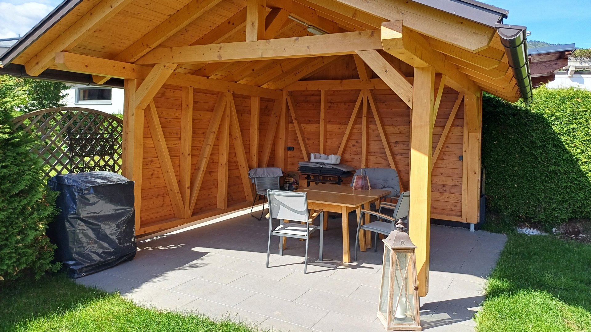 Holzgartenlaube mit Outdoor-Möbeln Eine Holzgartenlaube in einem gepflegten Garten mit Sitzmöbeln und Rasenfläche für Entspannung im Freien.